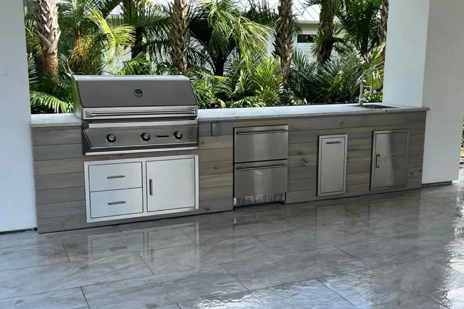 Linear outdoor kitchen installation with built-in grill, refrigerator drawers, and wood-plank cabinetry under covered patio by Jax Pavers in Jacksonville FL